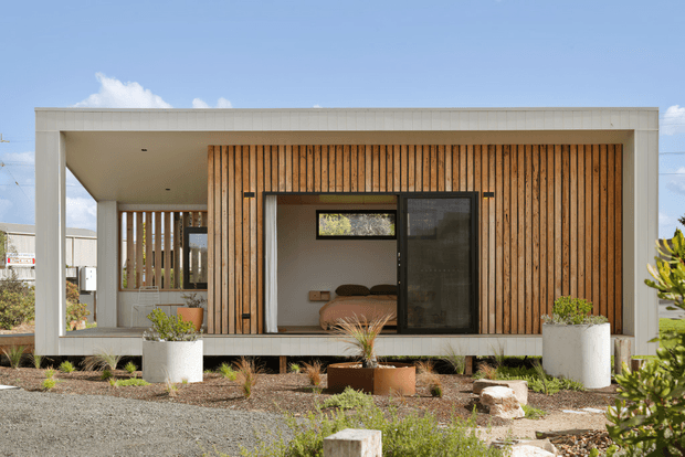 A compact modern single-storey dwelling with a flat-roofed rectangular form, white rendered frame, and a warm timber slat facade. Large glazed openings reveal a bedroom inside, while landscaped gravel planting beds and raised planters sit in front of the building.