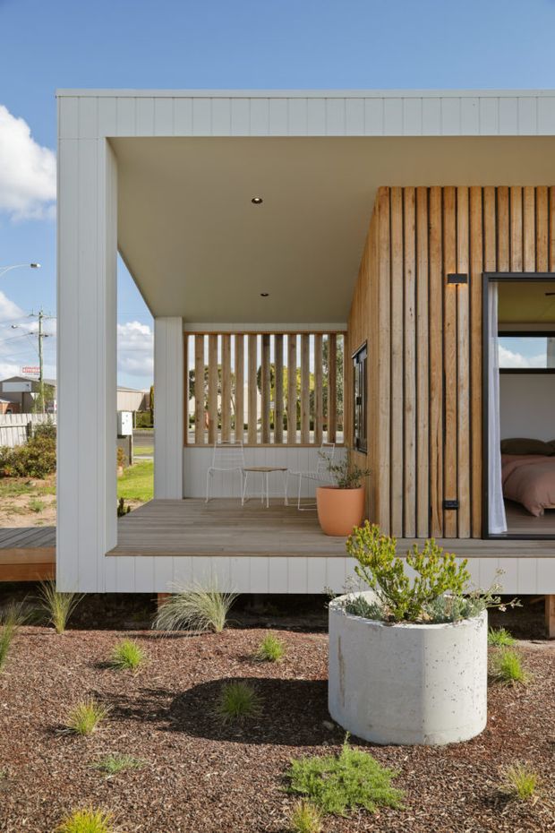 Exterior view of a compact contemporary home with a covered timber-deck porch, white vertical cladding, warm vertical timber screening, and a bedroom visible through an open glazed door. The home is raised slightly above a low-maintenance landscaped garden with native grasses and a large concrete planter.