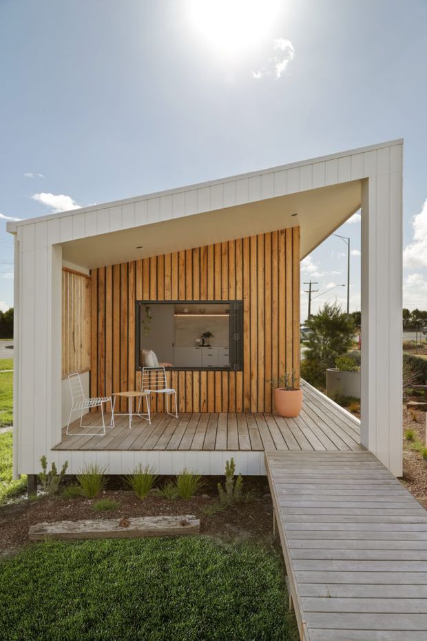A compact modern pavilion or small dwelling set within a landscaped outdoor setting. The design features a bold white rectangular frame, warm vertical timber wall cladding, a covered timber deck, and a raised timber walkway leading to the entrance. Outdoor chairs and a small table sit on the deck, with a glimpse of a simple interior kitchen through the opening. The composition is bright and contemporary, with strong geometric lines and a prominent skillion-style sloping roof form.