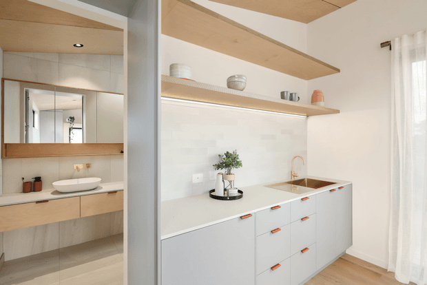 Bright, compact open-plan kitchen viewed beside a bathroom vanity. The kitchen features pale grey fixed cabinetry with small timber-toned handles, a light engineered-looking benchtop, white tiled splashback, timber floating shelves, and a timber-lined ceiling detail. A timber-framed bathroom vanity with a vessel basin and mirrored cabinet is visible through the opening on the left. Soft sheer curtains and light timber flooring contribute to an airy, warm, minimalist interior.