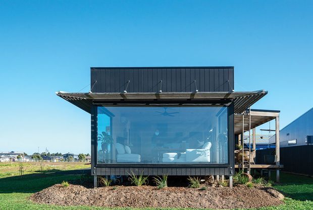 Front exterior view of a compact contemporary pavilion-like building with dark vertical cladding, a broad low-pitched roof with deep overhangs, and a full-width glazed living space. A covered side structure and open landscaped site are visible against a clear blue sky.