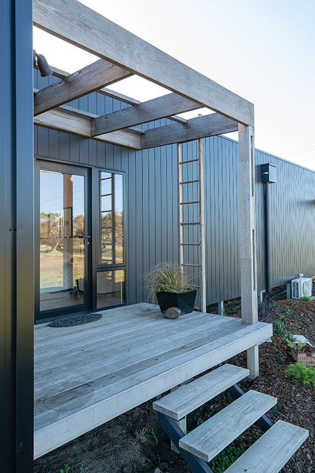 Exterior view of a compact contemporary building entrance with a raised timber deck and steps, dark vertical corrugated metal cladding, black-framed glazed doors, and a timber pergola-like overhead structure. A ladder, planter, outdoor lighting, and air-conditioning unit are visible, with a rural landscape reflected through the glazing.