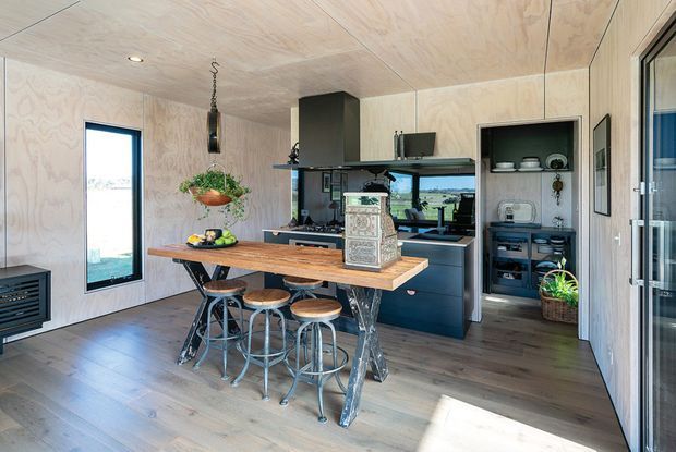 Compact open-plan kitchen with a timber island bench and industrial-style stools, dark blue-black cabinetry, integrated appliances, open shelving, and pale plywood wall and ceiling linings. Large glazing provides bright natural light and views of the surrounding landscape. The space has a warm, functional, contemporary cabin-like character with exposed timber finishes and metal accents.