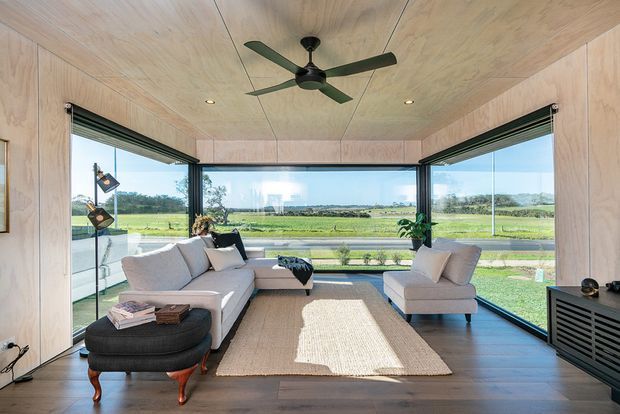 A bright contemporary living room enclosed by expansive corner glazing, overlooking open green fields. The room features light timber wall and ceiling lining, dark timber flooring, a pale sectional sofa, lounge chair, woven rug, black ceiling fan, and low dark media cabinetry. Retractable or folding glazed openings create a strong connection to the landscape.