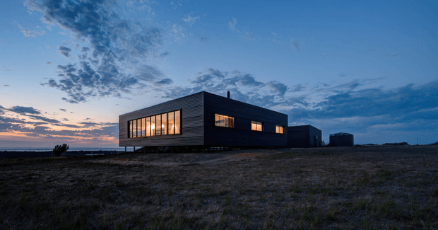 A low, modern rectangular house set in a sparse open landscape at dusk. The dark horizontal cladding, flat roof, long bands of warm-lit windows, and raised form create a strong contrast against the blue evening sky and distant horizon.