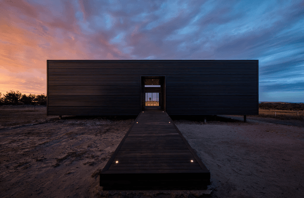 A minimalist single-storey rectangular building viewed from the front at dusk. The dark horizontally ribbed facade has a centrally positioned glazed entry, reached by a long timber walkway with small integrated lights. The building sits in a sparse, flat landscape under a dramatic blue and warm orange sunset sky.