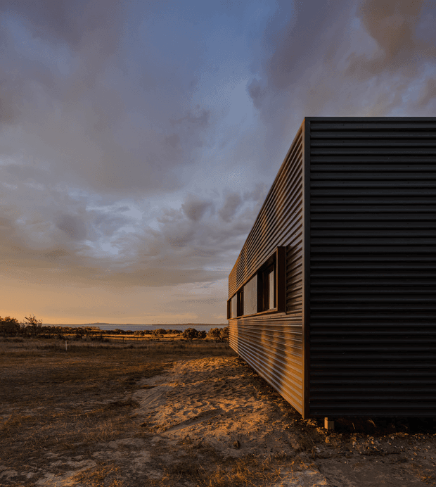 A small contemporary rectangular building viewed from the side at dusk, set in an open rural landscape. The facade features dark horizontal corrugated cladding, a timber-toned side wall with narrow horizontal boards, and a row of recessed windows. Dramatic cloud cover and warm low-angle light create a moody architectural exterior photograph.