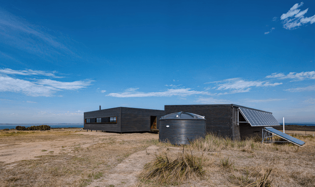 A low, dark-clad contemporary building set in a sparse coastal or rural landscape, photographed from outside. The structure has a long horizontal form, corrugated or ribbed dark exterior walls, large horizontal windows, a prominent cylindrical rainwater tank, and roof-mounted solar panels. Sandy ground and dune-like grasses surround the building under a bright blue sky.