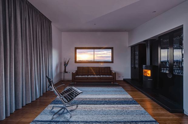 A refined contemporary living room with full-height grey curtains, warm timber flooring, a brown leather sofa, a black leather lounge chair, and a large layered blue-grey rug. A wide horizontal window frames a sunset view, while a black built-in fireplace and glazed display cabinetry create a strong feature wall. The room has a calm, moody atmosphere with warm firelight and understated furnishings.