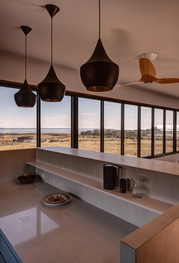 A warm, minimalist kitchen or open-plan living space with a long pale stone island and integrated raised shelving, surrounded by floor-to-ceiling windows overlooking a dry rural landscape and distant water. Several sculptural dark pendant lights hang from the ceiling, while a timber ceiling fan and sparse decorative objects add contrast. The interior has a refined, earthy palette and strong connection to the landscape.