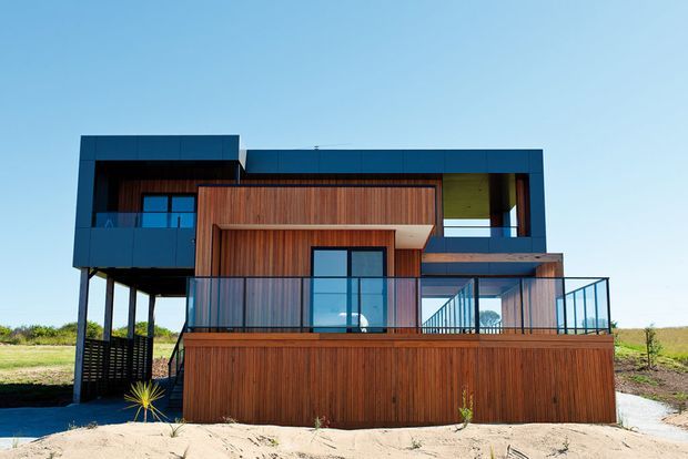 Exterior view of a contemporary multi-level house on a sandy, open coastal-looking site. The facade combines dark charcoal cladding and framing with extensive vertical timber screening and decking, large glazed openings, projecting upper volumes, and a glass-balustraded terrace. The elevated structure and open lower level are clearly visible.