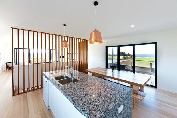 Bright open-plan kitchen with a large speckled stone island and integrated double sink, white cabinetry, light timber flooring, and warm timber vertical screening separating the kitchen from the adjoining dining area. Copper pendant lights hang above the island, while black-framed glazing provides views toward a green outdoor landscape.