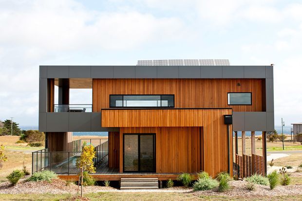 Modern two-storey coastal residence with a strong rectangular form, dark grey upper-level framing, and warm vertical timber cladding. The facade includes large glazed openings, a recessed balcony, external stairs, and a flat roof with rooftop solar panels. Sparse landscaping and glimpses of the sea reinforce the coastal setting.