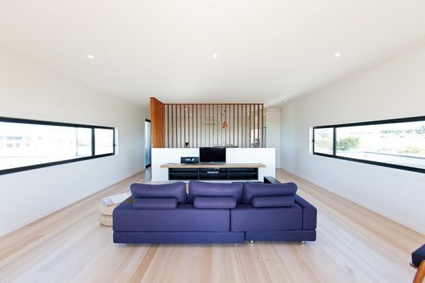 Bright, spacious contemporary living room with a large deep-blue sectional sofa facing a wall-mounted television and low white media console. Pale timber flooring, white walls and ceiling, expansive horizontal black-framed windows, and a vertical timber slat screen create a clean, open interior.