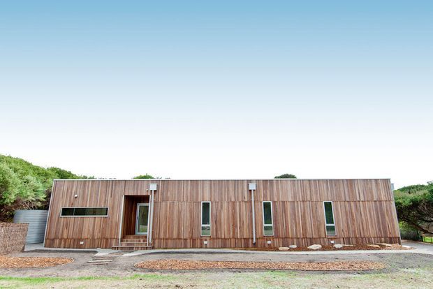 Single-storey contemporary building with a long, low horizontal form and a facade clad in vertical timber boards. Narrow vertical windows and a glazed entry door punctuate the facade, with a simple flat or very low-pitched roofline. The building sits in a sparse landscaped setting with gravel paths and low vegetation.