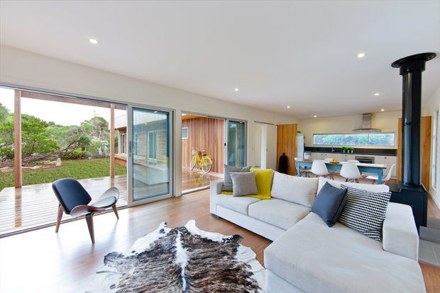 Bright open-plan living room with a large white sectional sofa, mid-century-style lounge chair, cowhide rug, and black freestanding wood stove. Full-height glazing opens toward a landscaped outdoor area, while the dining and kitchen zones are visible beyond. The room has white walls and ceiling, light timber flooring, timber and glass doors, and a warm contemporary fit-out.