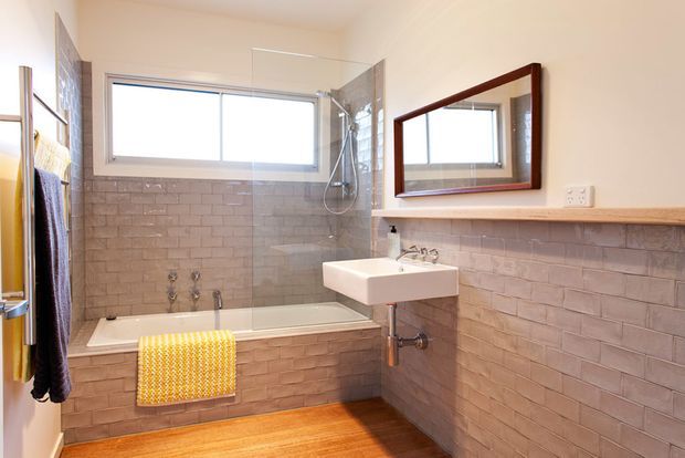 Bright, compact bathroom with a white bathtub and wall-mounted shower behind a clear glass screen, a white pedestal-style basin, exposed plumbing, grey-beige subway tile wainscoting, white upper walls, a timber shelf, and a dark timber-framed rectangular mirror. A warm timber floor and colourful towels add contrast.
