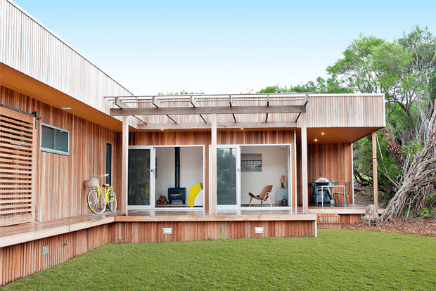 Exterior view of a low-rise contemporary timber-clad home opening onto a broad lawn. The facade features vertical wood cladding, extensive sliding glass doors, a raised timber deck, pergola-like roof structure, sheltered outdoor sitting area, bicycles, and built-in exterior screening. Mature vegetation frames the right side of the property.