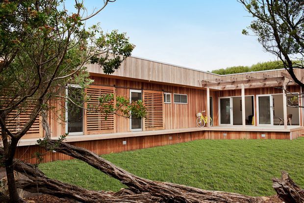 Single-storey contemporary timber-clad residence viewed from a landscaped garden. The building has a low, horizontal form with warm vertical timber cladding, timber slatted screening, a partially covered deck or verandah, and extensive glazed doors and windows opening toward the lawn. Mature trees frame the foreground.