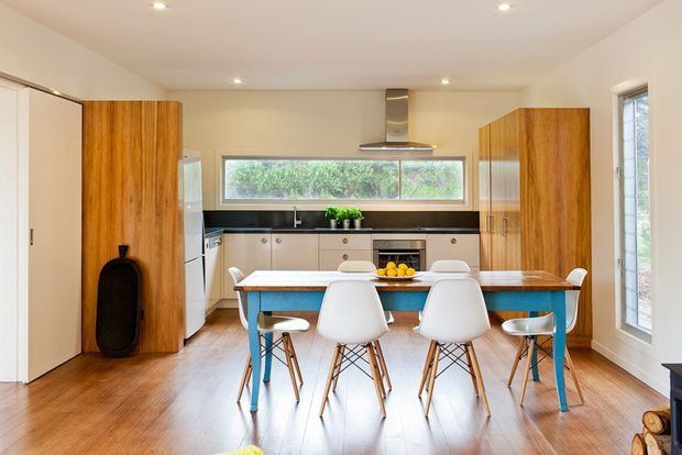 Bright, open-plan kitchen and dining area with warm timber flooring, white walls and cabinetry, tall timber joinery, a dark benchtop and splashback, and a colourful turquoise dining table surrounded by modern white chairs. A long horizontal window provides natural light above the kitchen work zone, with additional glazing at the side.