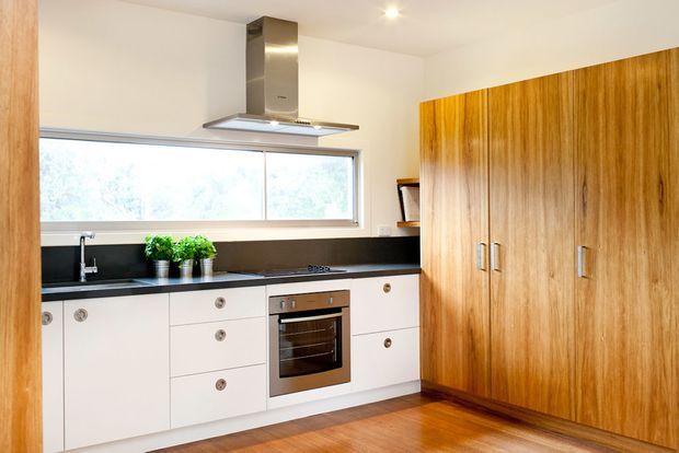 Bright contemporary kitchen with white handleless cabinetry, dark benchtops and splashback, integrated oven, stainless-steel rangehood, timber flooring, and a large floor-to-ceiling timber storage wall. A wide horizontal window provides natural light and views to greenery.