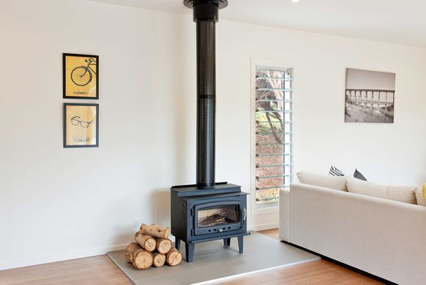 Bright, compact living room with a freestanding black wood-burning stove and flue as the focal point. The room has white walls, timber flooring, a light upholstered sofa, a small stack of firewood, framed wall art, and a louvred window providing natural light.
