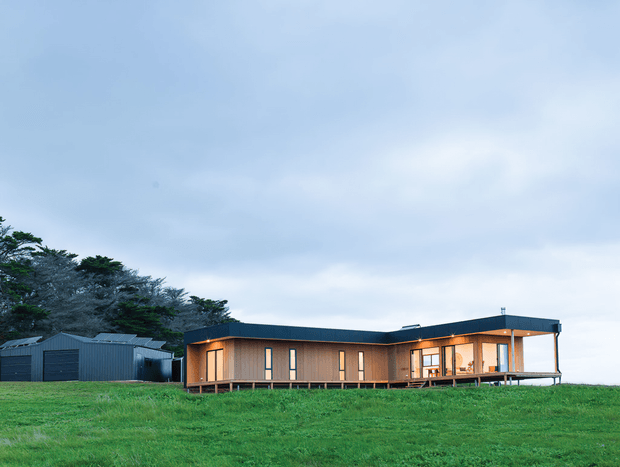 A low, elongated single-storey contemporary house set on a broad grassy site, with dark metal-clad walls and roof, extensive warm timber-framed glazing, and a covered verandah or deck. A detached dark-clad outbuilding sits to the left, with trees behind and an overcast sky.