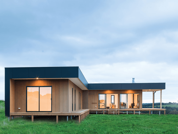 A modern single-storey pavilion-style house set in a grassy rural landscape. The building combines dark blue-black flat roof planes with warm vertical timber wall cladding, extensive floor-to-ceiling glazing, and a broad elevated timber deck and covered outdoor living area. The low horizontal form and open site are prominent, with warm interior lighting visible through the windows.