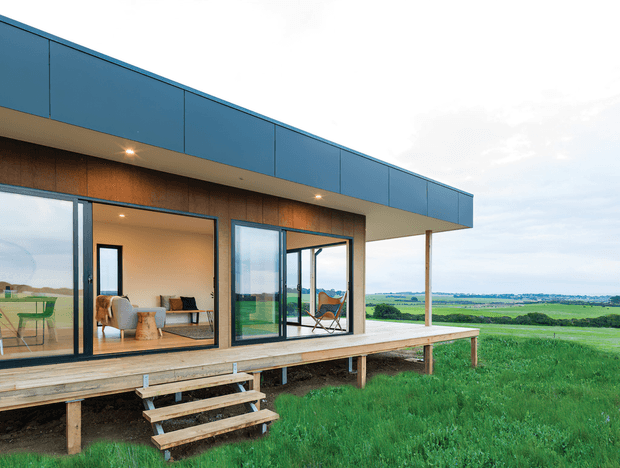 A minimalist single-storey modern pavilion-style house set in an open rural landscape. The building has a long flat roof with blue metal fascia, warm timber-clad walls, extensive floor-to-ceiling glazing, and a raised timber deck with steps. The interior and deck are visible through the open glass doors, with expansive green fields beyond.