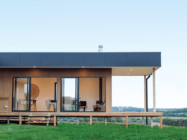 A modern single-storey pavilion-style house elevated slightly above a grassy site, with a flat roof, dark metal roof fascia, extensive floor-to-ceiling glazing, and a covered timber deck. The open-plan interior is visible through the glass, with simple furnishings and warm-toned wall finishes. Rolling rural landscape is visible beyond the building.