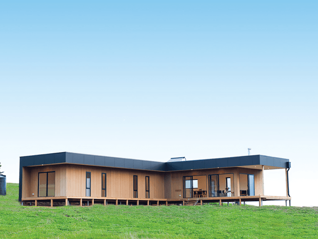 A single-storey contemporary house set on a gently sloping grassy site. The low, elongated building has warm vertical timber cladding, dark flat roof sections, extensive dark-framed glazing, and a raised timber deck wrapping around the living areas. The image is a clear exterior architectural view under a bright blue sky.