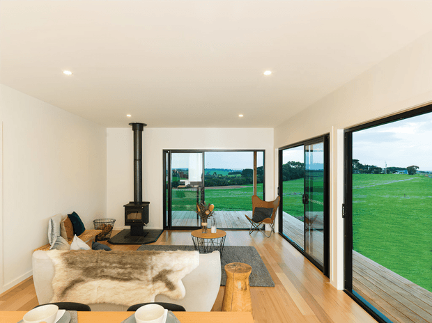 A bright, open-plan living room with pale walls, timber flooring, a white curved sofa draped with a sheepskin throw, a compact black wood-burning stove, and a timber coffee table. Large black-framed sliding glass doors open onto a timber deck and expansive green rural fields. The room has a simple contemporary interior with recessed ceiling lights and a warm, natural palette.