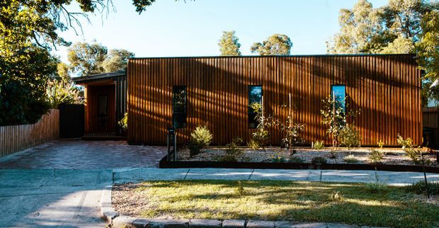 Single-storey contemporary residence with a long, low rectangular form and dark vertical timber-clad facade. Narrow vertical windows and a recessed entry punctuate the facade, while a paved driveway or forecourt and simple landscaped garden sit in front. Mature trees surround the suburban property.
