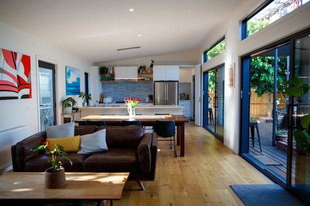 Bright open-plan living and kitchen interior with a dark leather sofa in the foreground, a timber coffee table, dining table and chairs, and a compact white kitchen with a central island. Wide black-framed glazed doors and windows open toward an outdoor deck, while colourful artwork, plants, timber flooring, and a patterned tiled splashback add visual interest.