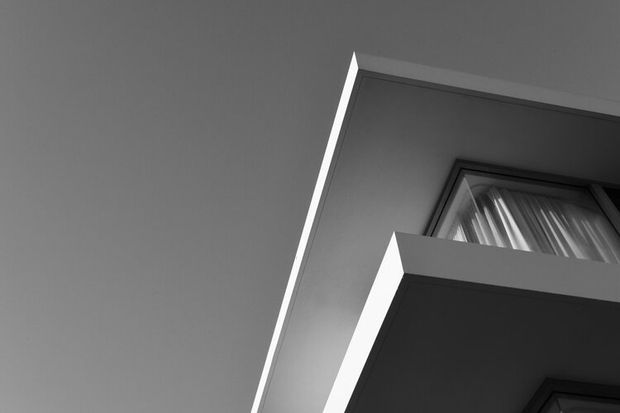 Black-and-white upward-looking architectural photograph focused on the sharp geometric edges of a modern building facade and projecting balcony or overhang. A partially visible window with curtains appears near the upper right, while the minimalist composition emphasizes angular planes, shadow, and contrast against the sky.