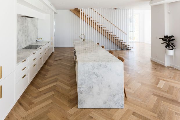 Bright open-plan kitchen and living area with white cabinetry, a large marble-look island, timber herringbone flooring, and a staircase with slim white balusters and timber treads. The space has abundant natural light and a clean, refined contemporary fit-out.
