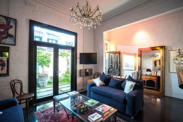 A richly styled living room in a heritage-style interior, featuring exposed painted brick walls, dark polished timber flooring, ornate ceiling mouldings, a crystal chandelier, tall black-framed glazed doors, a deep navy sofa, antique-style bentwood chairs, a glass coffee table, and large decorative mirrors. The room opens visually toward a warmly lit adjoining space.