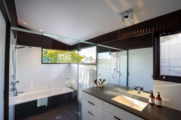 A contemporary bathroom with a large freestanding-style bathtub enclosed by frameless glass, a separate glass shower, and a double vanity with a dark stone benchtop and integrated basins. Dark vertical wall panelling, white wall tiles, charcoal floor tiles, mirrored surfaces, and clerestory-style glazing create a high-contrast, refined interior.