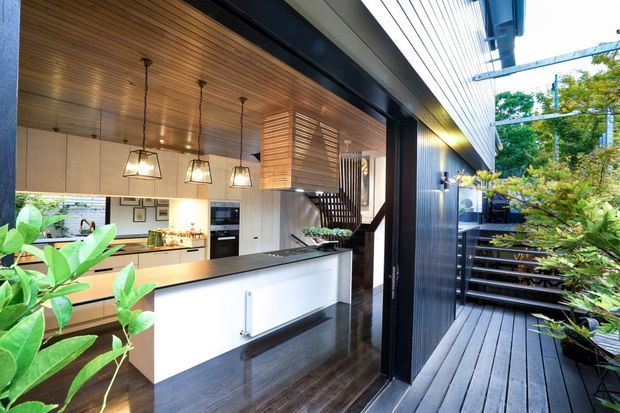 A contemporary open-plan kitchen viewed from an adjoining outdoor deck. The kitchen features a large white island with dark benchtop, pale cabinetry, integrated appliances, pendant lights, a timber-slatted ceiling, dark timber flooring, and a timber screen near the stair. Black-framed full-height glazing and dark exterior cladding connect the interior to a landscaped deck and garden.