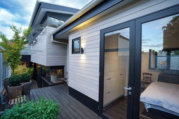 Exterior view of a contemporary multi-level home with horizontal light cladding, dark trim, and a timber deck walkway. Glazed doors reveal a bedroom inside, while landscaped planters and a recessed lower-level outdoor area are visible around the building.