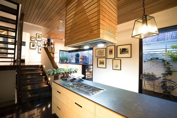 Contemporary open-plan kitchen viewed from the island, featuring pale timber cabinetry, a dark stone benchtop, and a large timber-lined rangehood enclosure integrated with the ceiling. Black open-riser stairs, extensive glazing, framed artwork, and a hanging geometric pendant contribute to the refined, warm interior. A small courtyard is visible through the windows.