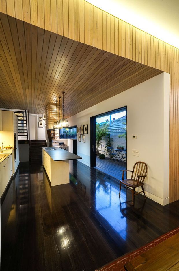 A narrow, open-plan contemporary interior combining a kitchen and living area. Dark polished timber flooring contrasts with a pale kitchen island and cabinetry, while extensive warm timber wall and ceiling panelling creates a continuous slatted canopy. Black-framed glazing opens to a small outdoor deck and courtyard. Pendant lights, framed artwork, a timber chair, and a stairway are visible.