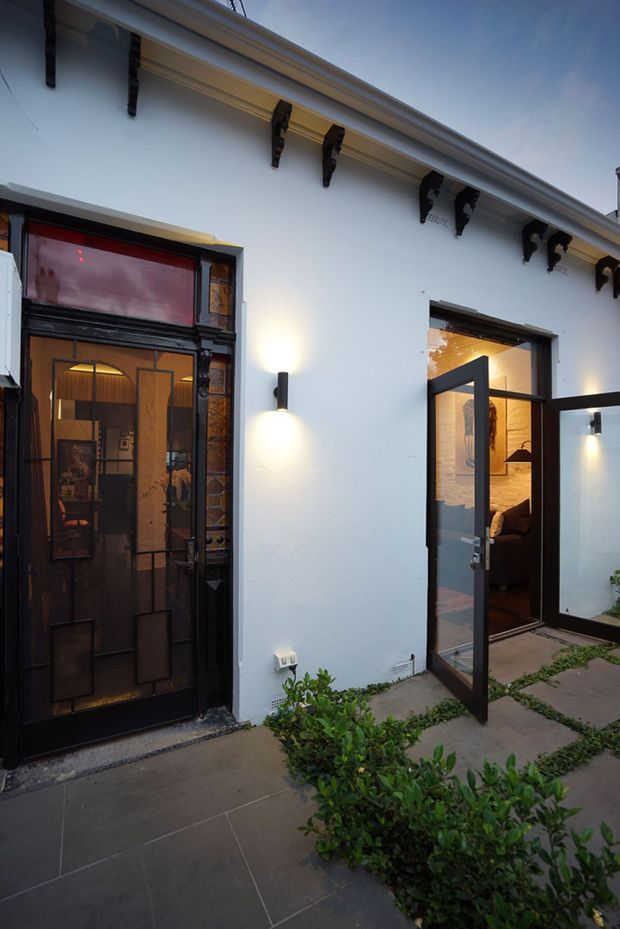 A dusk exterior view of a white rendered heritage-style building facade with ornate dark timber corbels beneath the eaves. A black decorative glazed entry door sits beside an open black-framed glass doorway revealing a warmly lit interior and staircase. Wall-mounted lights illuminate the facade, while a paved walkway and clipped greenery occupy the foreground.