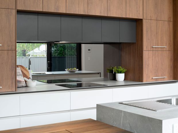 Contemporary kitchen with extensive warm timber cabinetry, sleek white lower drawers, dark grey overhead cabinetry, a pale stone benchtop, integrated black cooktop, and a substantial concrete-toned island with stainless-steel sink and draining board. A mirrored or reflective splashback visually enlarges the room and reflects the rear glazing and garden beyond. The space is clean-lined, functional, and softly natural in tone.