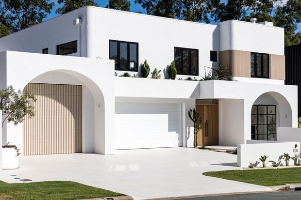 A contemporary two-storey white rendered residence with strong geometric forms, arched openings, flat rooflines, black-framed windows, timber-toned vertical garage screening, and a pale timber entry door. The front facade is landscaped with sculptural planting, low white walls, and a broad light-coloured driveway.
