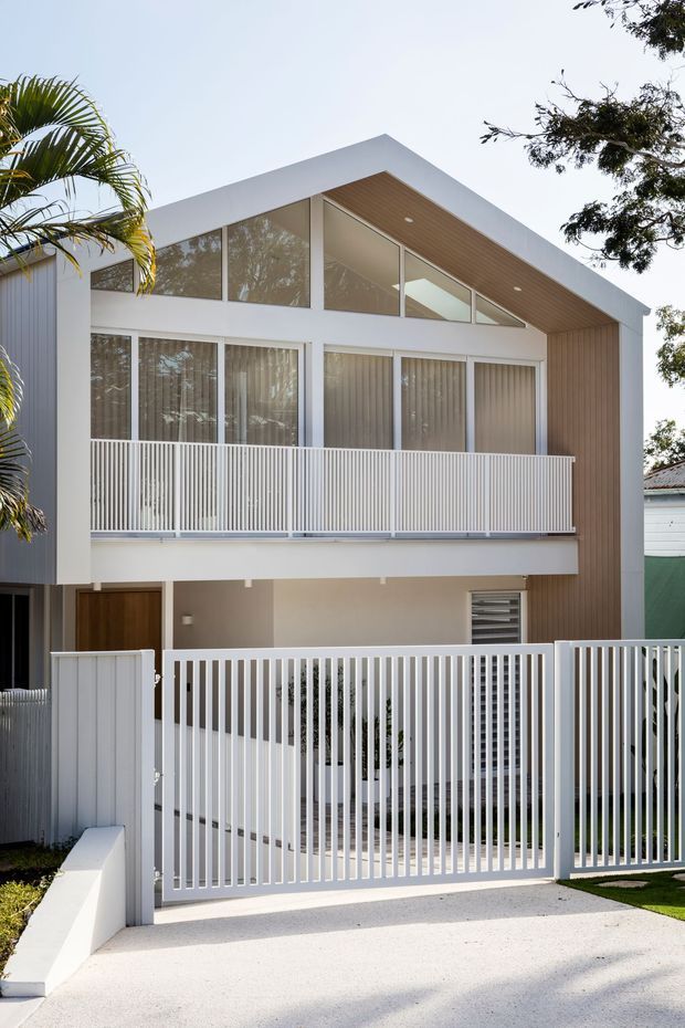 Front exterior view of a contemporary two-storey residence with a prominent pitched/gabled roof form, white rendered walls, extensive upper-level glazing, warm timber-lined soffit, and white vertical-slatted balcony and driveway fencing. The property includes a recessed entry and integrated garage or carport area, viewed from the driveway.