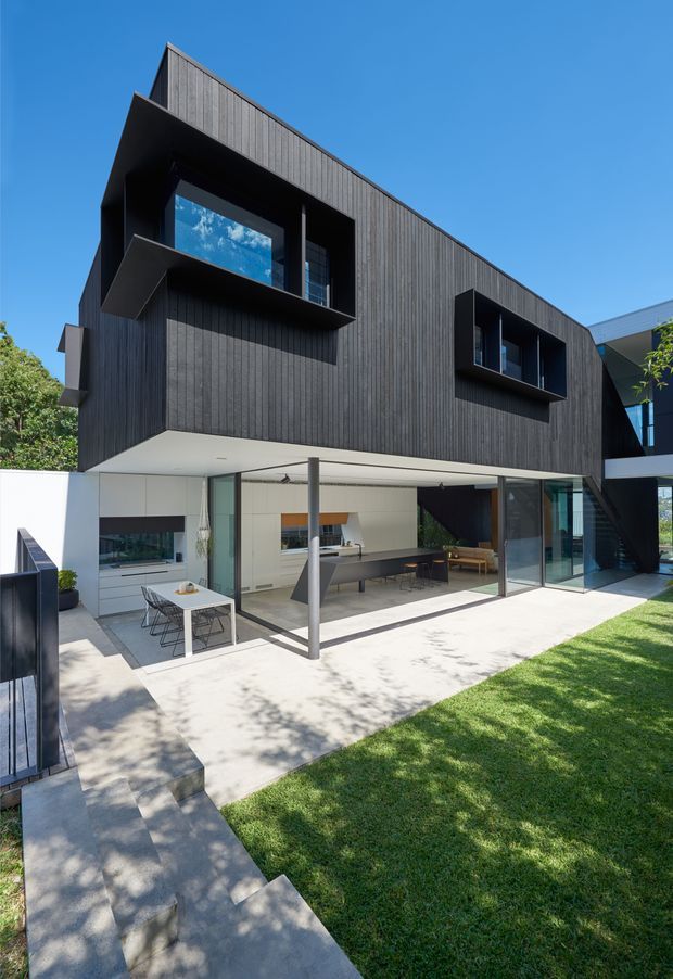 A contemporary multi-storey residential exterior viewed from a landscaped garden. The upper level is clad in dark vertical timber boards with projecting, deeply recessed window boxes. The ground floor is largely open and glazed, creating an indoor-outdoor living and dining area with a kitchen visible inside. A pale concrete terrace, outdoor dining table, lawn, steps, and a dark metal boundary gate complete the scene. The design has strong cantilevered forms, clean geometric lines, and extensive sliding glass doors.