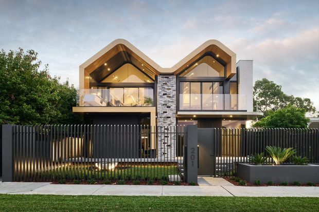 Contemporary multi-storey residential facade photographed from the street, featuring two prominent folded gable-like roof forms, warm timber soffits, extensive upper-level glazing, dark vertical metal fencing, and a central stacked-stone feature wall. Landscaped planting and a numbered entry gate sit in front of the house.