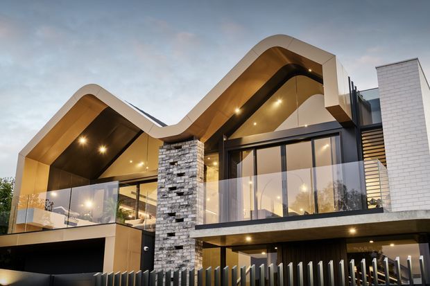Contemporary multi-level residential facade at dusk, featuring two dramatic curving pitched roof forms with warm timber-toned soffits, extensive glazing, balconies, a prominent stacked-stone column, white brick cladding, and a dark vertical-slat fence and garage frontage.