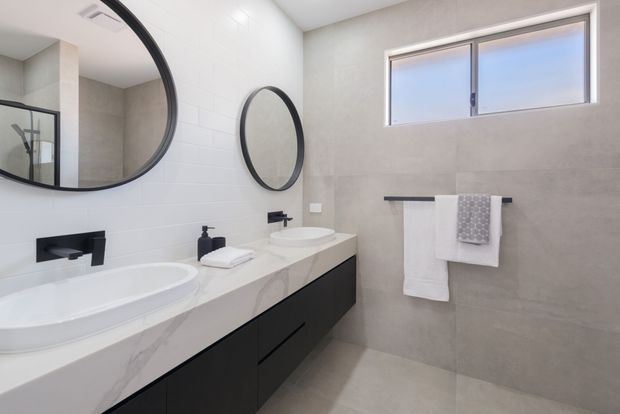 Contemporary bathroom with a double vanity, white vessel basins, a white marble-look benchtop, matte black wall-mounted fixtures and dark cabinetry. Large round mirrors, pale grey wall and floor tiles, a frameless shower enclosure, and a horizontal frosted window create a clean, refined space.
